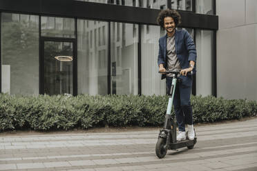 Lächelnder junger Mann, der einen elektrischen Roller auf dem Fußweg vor einem modernen Gebäude fährt - MFF08954