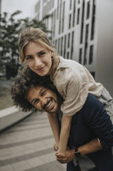 Happy young man giving piggyback ride to girlfriend in front of modern building - MFF08974