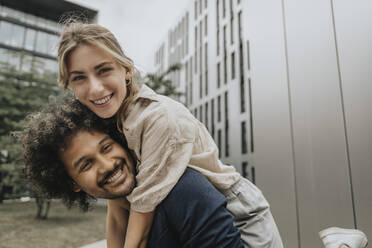 Smiling young man piggybacking girlfriend in front of modern building - MFF08975