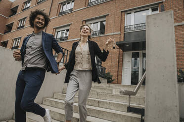 Happy couple holding hands walking down stairs of apartment building - MFF08980