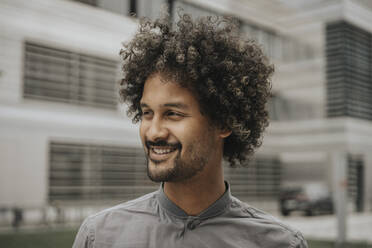 Lächelnder junger Mann mit lockigem Haar vor einem modernen Gebäude - MFF08987