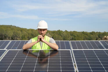 Ingenieur mit Schutzhelm steht mit gefalteten Händen zwischen Solarzellen auf dem Dach - VEGF05530