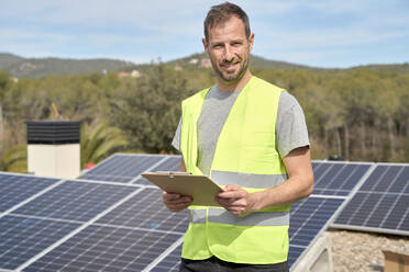 Lächelnder Ingenieur mit Bauplan vor Sonnenkollektoren an einem sonnigen Tag - VEGF05535