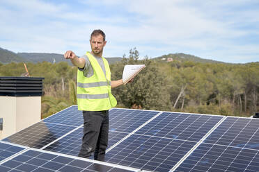 Ingenieur mit Bauplan inmitten von Solarzellen auf dem Dach - VEGF05538