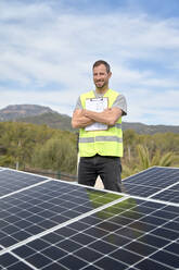 Lächelnder Ingenieur mit verschränkten Armen, der mit einem Bauplan vor einem Solarpanel steht - VEGF05540