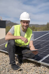 Lächelnder Ingenieur mit Tablet-PC, der an einem sonnigen Tag bei Sonnenkollektoren hockt - VEGF05569