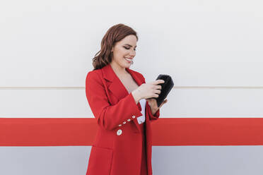 Happy businesswoman using mobile phone standing in front of wall - JRVF02892