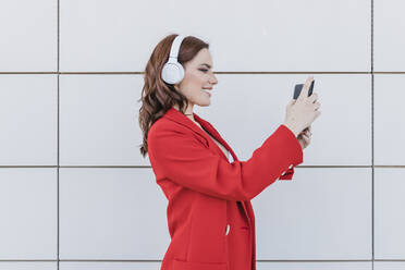 Glückliche Geschäftsfrau mit drahtlosen Kopfhörern und Smartphone an der Wand - JRVF02896