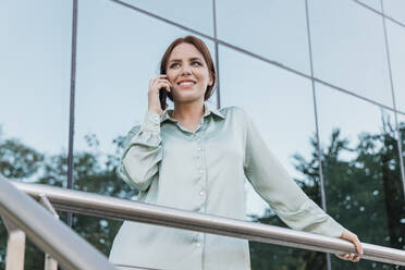 Lächelnde Geschäftsfrau, die vor einem modernen Gebäude mit einem Mobiltelefon spricht - JRVF02904