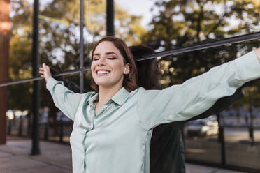 Smiling businesswoman with arms outstretched leaning on glass wall - JRVF02909