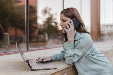 Lächelnde Geschäftsfrau, die einen Laptop benutzt und mit einem Smartphone telefoniert - JRVF02911