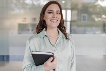 Happy businesswoman with diary standing in front of glass window - JRVF02913