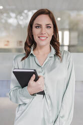 Smiling businesswoman with diary standing in front of glass window - JRVF02914