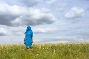 Woman covered with blue scarf standing amidst grass at meadow - OJF00497