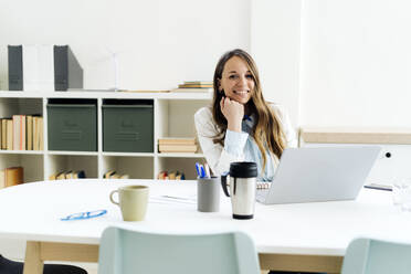 Smiling businesswoman with laptop sitting hand on chin at desk in office - GIOF14964