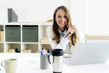 Happy businesswoman holding pen sitting with laptop at desk - GIOF14965