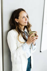 Geschäftsfrau mit Kaffeetasse an der Wand lehnend im Büro - GIOF14977