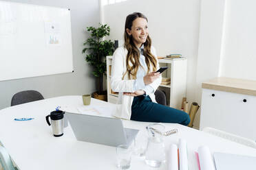 Smiling businesswoman holding smart phone sitting at desk in office - GIOF14987