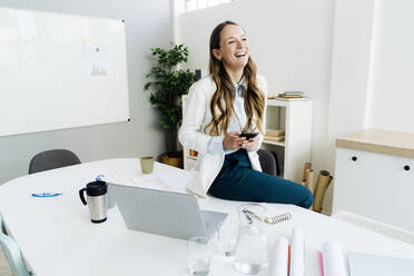 Happy businesswoman holding smart phone sitting on desk in office - GIOF14988