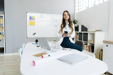 Businesswoman holding smart phone sitting on desk in office - GIOF14989