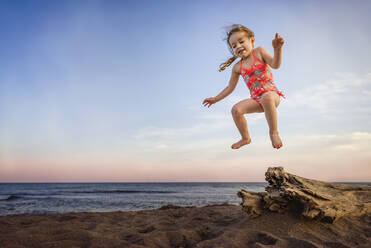 Kleines Mädchen springt von Treibholz am Strand im Sommer - CAVF96005