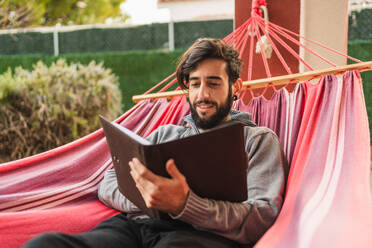 Entspannter Hipster-Mann in Hängematte beim Lesen eines Buches - CAVF96026