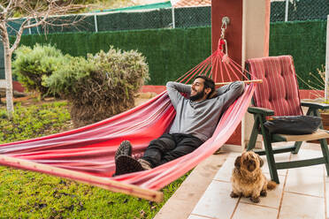 Entspannter Mann, der in einer Hängematte liegt und sich zu Hause mit seinem Hund vergnügt - CAVF96031