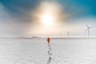 A boy running through snow during sunrise on a wind farm - CAVF96050