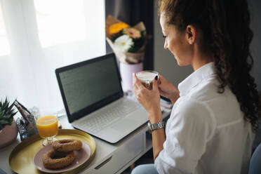 Frau schaut auf ihren Laptop und lächelt, während sie eine Tasse Kaffee in der Hand hält - CAVF96055