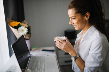 Frau schaut auf ihren Laptop und lächelt, während sie eine Tasse Kaffee in der Hand hält - CAVF96058