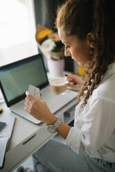 Frau sitzt an ihrem Schreibtisch und schaut auf ihre Kreditkarte - CAVF96060