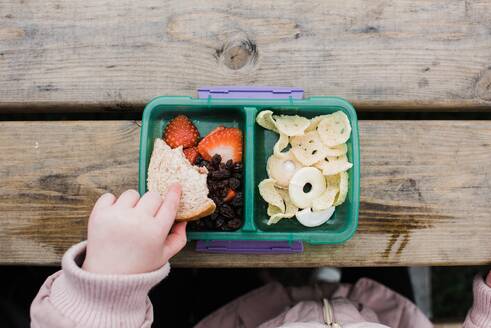 Kids hand eating lunch from a lunch box outside on a picnic table - CAVF96083