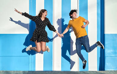 Full body of active positive couple holding hands while jumping near striped wall on sunny street in city on summer day - ADSF34163