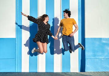 Ganzkörperansicht eines aktiven positiven Paares, das sich an den Händen hält, während es in der Nähe einer gestreiften Wand auf einer sonnigen Straße in der Stadt an einem Sommertag springt - ADSF34164