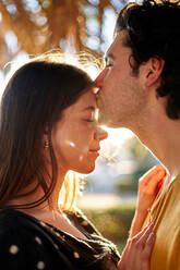 Side view of romantic man with closed eyes kissing forehead of girlfriend while standing on sunny street against blurred background - ADSF34165