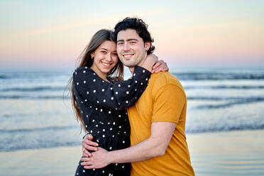 Verliebtes Paar in Freizeitkleidung, das sich umarmt und zärtlich streichelt, während es an einem Sommerabend am Sandstrand steht - ADSF34167