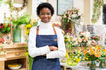 Smiling African American female florist in apron looking at camera while standing among colorful bouquets of flowers in floral shop - ADSF34177