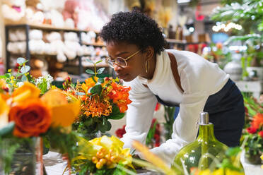 Seitenansicht einer afroamerikanischen Floristin mit geschlossenen Augen, die einen Blumenstrauß mit grünen Zweigen in einer Vase genießt, während sie in einem Blumenladen steht - ADSF34178