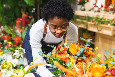 Afroamerikanische Blumenhändlerin mit geschlossenen Augen, die einen Blumenstrauß mit grünen Zweigen in einer Vase genießt, während sie in einem Blumenladen steht - ADSF34179