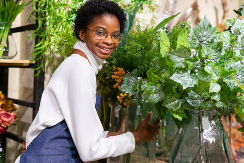 Positive afroamerikanische Floristin, die in die Kamera schaut, während sie in der Nähe von grünen Tolmiea-Pflanzen bei der Arbeit in einem modernen Blumengeschäft steht - ADSF34180