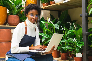 Seitenansicht einer konzentrierten afroamerikanischen Floristin, die auf einem Laptop tippt, während sie in einem Blumenladen mit einer Fülle von Blumen sitzt - ADSF34184