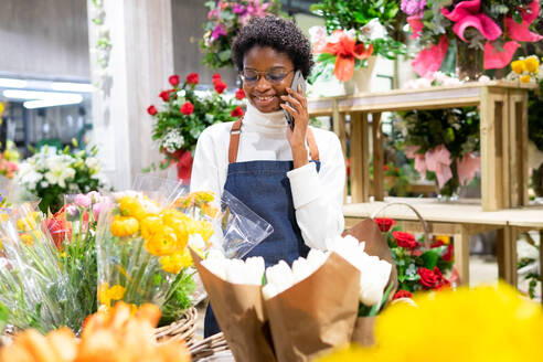 Positive afroamerikanische Floristin in Schürze, die mit einem Smartphone spricht, während sie in einem Blumenladen mit verschiedenen Blumensträußen steht - ADSF34185