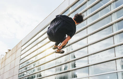 From below back view of anonymous male in casual clothes jumping and performing parkour trick in air on urban street with modern buildings - ADSF34186