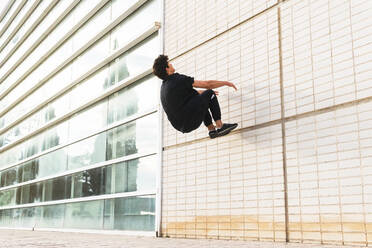 Seitenansicht eines Mannes in voller Länge in schwarzem Outfit, der einen akrobatischen Parkour an einer Gebäudewand ausführt - ADSF34188