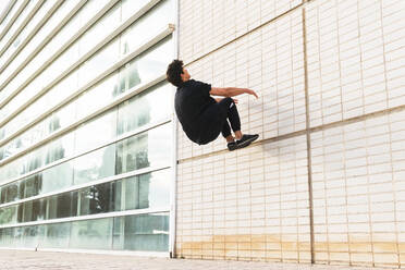 Side view of full length man in black outfit performing acrobatic parkour on wall of building - ADSF34188