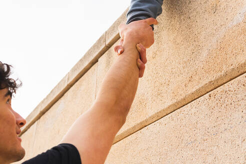 Seitenansicht einer anonymen Person in grauer Kleidung auf dem Dach, die die Hand eines Mannes in einer starken Umklammerung auf der Außenseite hält - ADSF34190