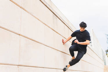 Seitenansicht eines Mannes in voller Länge in schwarzem Outfit, der einen akrobatischen Parkour-Stunt ausführt, während er über eine Gebäudewand springt - ADSF34192