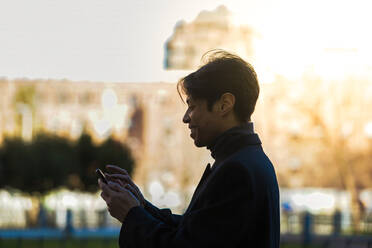 Seitenansicht eines lächelnden asiatischen Mannes im formellen schwarzen Anzug, der auf der Straße spazieren geht und Nachrichten auf seinem Smartphone liest - ADSF34193
