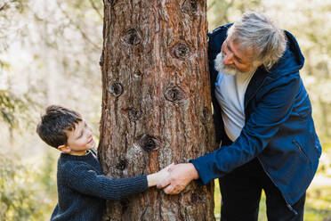 Side view of cute little boy and aged man with gray hair standing in forest around wood and holding hands on blurred background - ADSF34225
