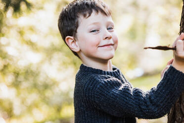 Side view of cheerful boy in warm sweater tears bark from tree and looking at camera while standing in autumn forest on sunny day on blurred background - ADSF34228